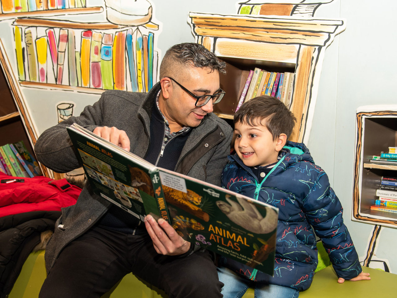 A man and a little boy sat looking at a large book together. They are both smiling and the man is pointing to something on the page
